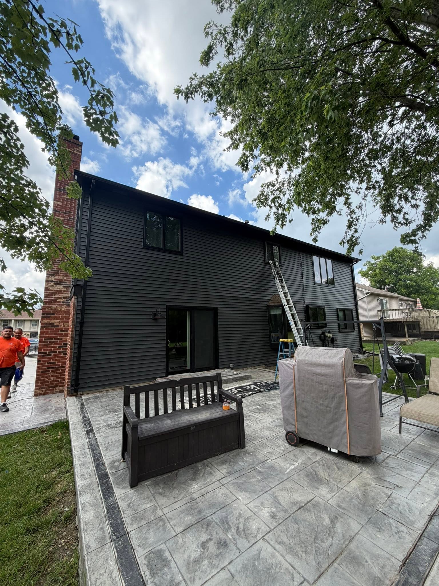 Rear of a two-story home painted deep charcoal with black window trim, a stamped concrete patio, and Brake crew members working