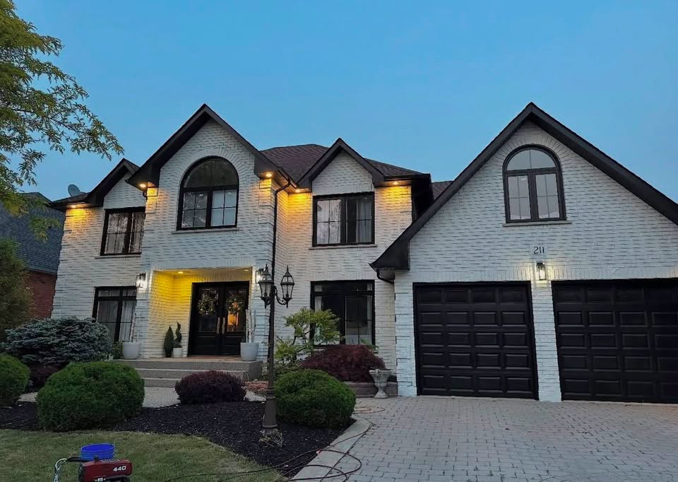 A large two-story brick home in Metro Detroit repainted in cream with dark trim and black garage doors, lit warmly at dusk